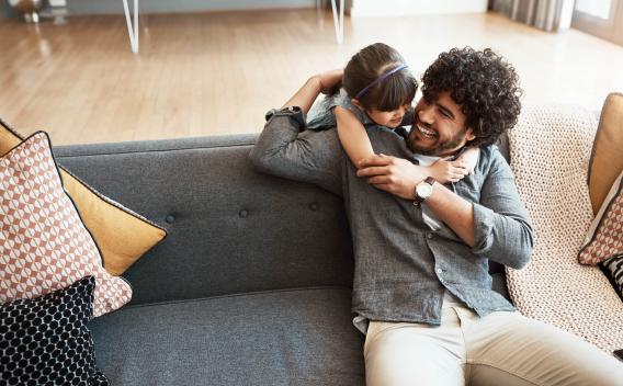 A father and daughter share a joyful moment, hugging and laughing on a modern grey sofa in a bright living room.
