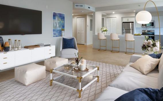 A bright and modern living room with white and gold decor, a mounted TV, and a view into an open-concept kitchen with a bar area.