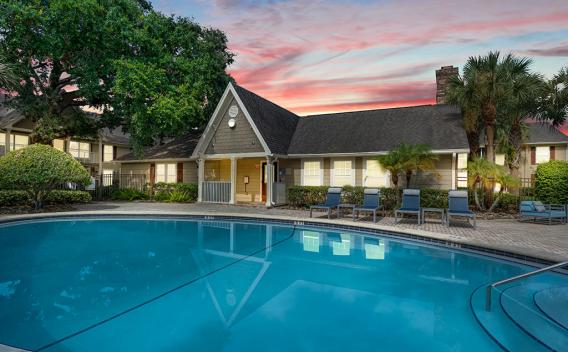 A serene pool area with lounge chairs and lush landscaping, set against a vibrant sunset sky and a cozy clubhouse.
