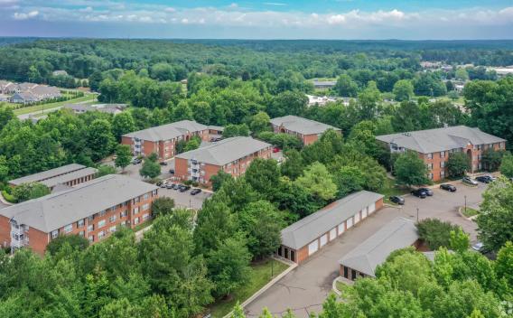 Aerial view of Heritage Apartments showcasing brick apartment buildings surrounded by lush green trees, with ample parking and nearby road access.