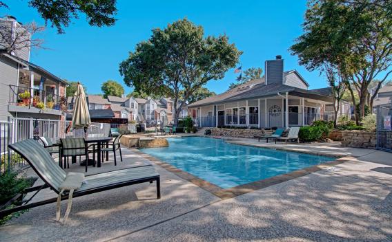 Sparkling resort-style swimming pool surrounded by lounge chairs, umbrellas, and lush landscaping with the clubhouse in the background.
