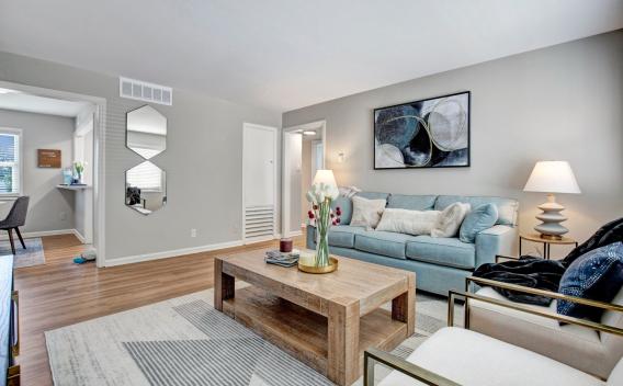 A stylish and cozy living room at Alcove at Seahurst, featuring a soft blue sofa adorned with decorative pillows, a rustic wooden coffee table with a floral centerpiece, and two modern accent chairs with gold metal frames. The space is illuminated by warm table lamps, and abstract wall art enhances the contemporary decor. An open doorway leads to a small dining area, and large windows bring in natural light, creating an inviting atmosphere. ​​