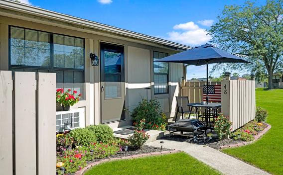 Private patio with umbrella and outdoor seating, surrounded by blooming flowers and well-manicured lawn outside apartment unit 48