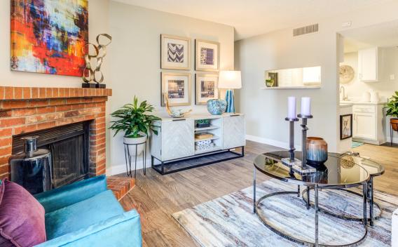 A stylish and contemporary living room at Santana Ridge featuring a cozy brick fireplace with vibrant abstract artwork above it. The room is furnished with a teal accent chair, a modern white console table adorned with decorative pieces, and a sleek glass coffee table with candles. The open-concept design leads to a dining area and kitchen with white cabinetry, creating a bright and inviting space. Warm wood flooring and indoor plants add a touch of comfort and elegance.