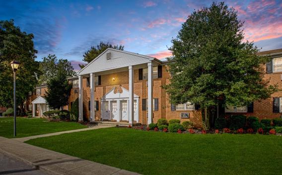 A grand entrance to Silver Lake Manor Apartments at twilight, showcasing a stately red brick building with tall white columns and an elegant gabled roof. The manicured lawn and vibrant flower beds add to the curb appeal, while a lamppost lights up the scenic sidewalk leading to the entrance. The sunset sky in the background creates a warm, inviting ambiance. ​​