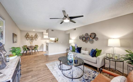 A modern open-concept living space at Skyline Apartments featuring a stylish living room, dining area, and kitchen. The living room is furnished with a gray sofa adorned with blue and green accent pillows, a round glass coffee table, and a leather accent chair. Decorative wall art, table lamps, and lush indoor plants enhance the ambiance. The dining area includes a wooden table with four green velvet chairs, while the kitchen features white cabinetry and a pass-through counter, creating a seamless and invit