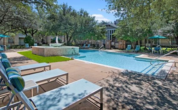 A luxurious outdoor pool area in a residential community, surrounded by mature trees and lush landscaping. The pool features a built-in water fountain, lounge chairs with blue and green striped pillows, and patio tables with teal umbrellas. A modern clubhouse with large windows is visible in the background, adding to the relaxing, resort-like atmosphere. ​​