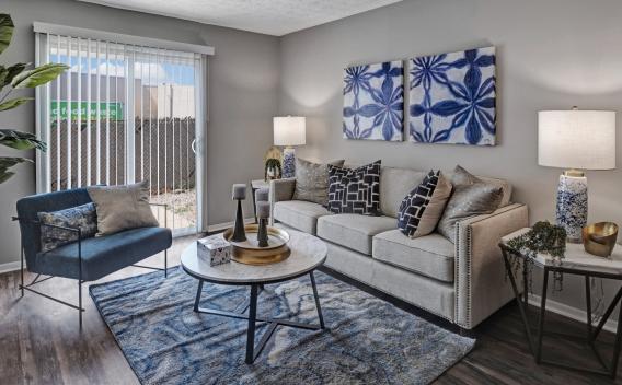 A stylish living room at Wilson Plaza featuring a beige fabric sofa adorned with decorative throw pillows, a navy blue accent chair, and a marble coffee table with a gold tray centerpiece. The space is illuminated by modern table lamps, and the decor includes blue abstract wall art and a patterned area rug. A sliding glass door with vertical blinds leads to an outdoor patio area, bringing in natural light. ​​