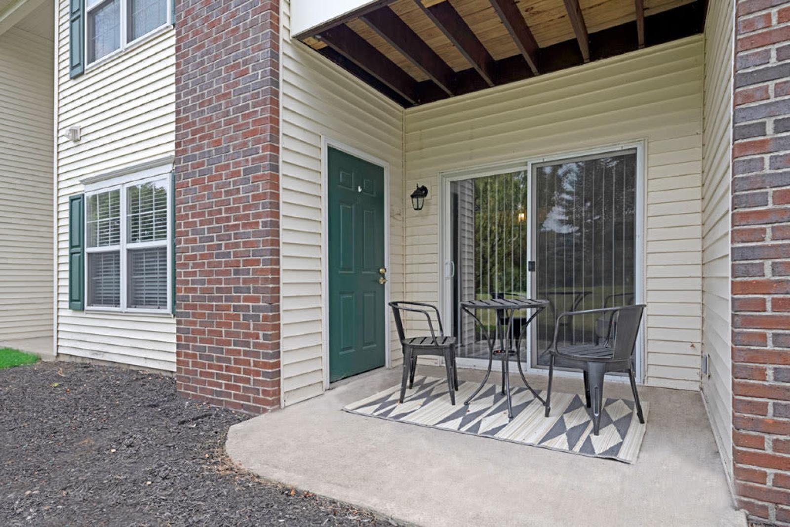 A cozy outdoor patio space with a small table, two chairs, and a patterned rug, overlooking a green yard.