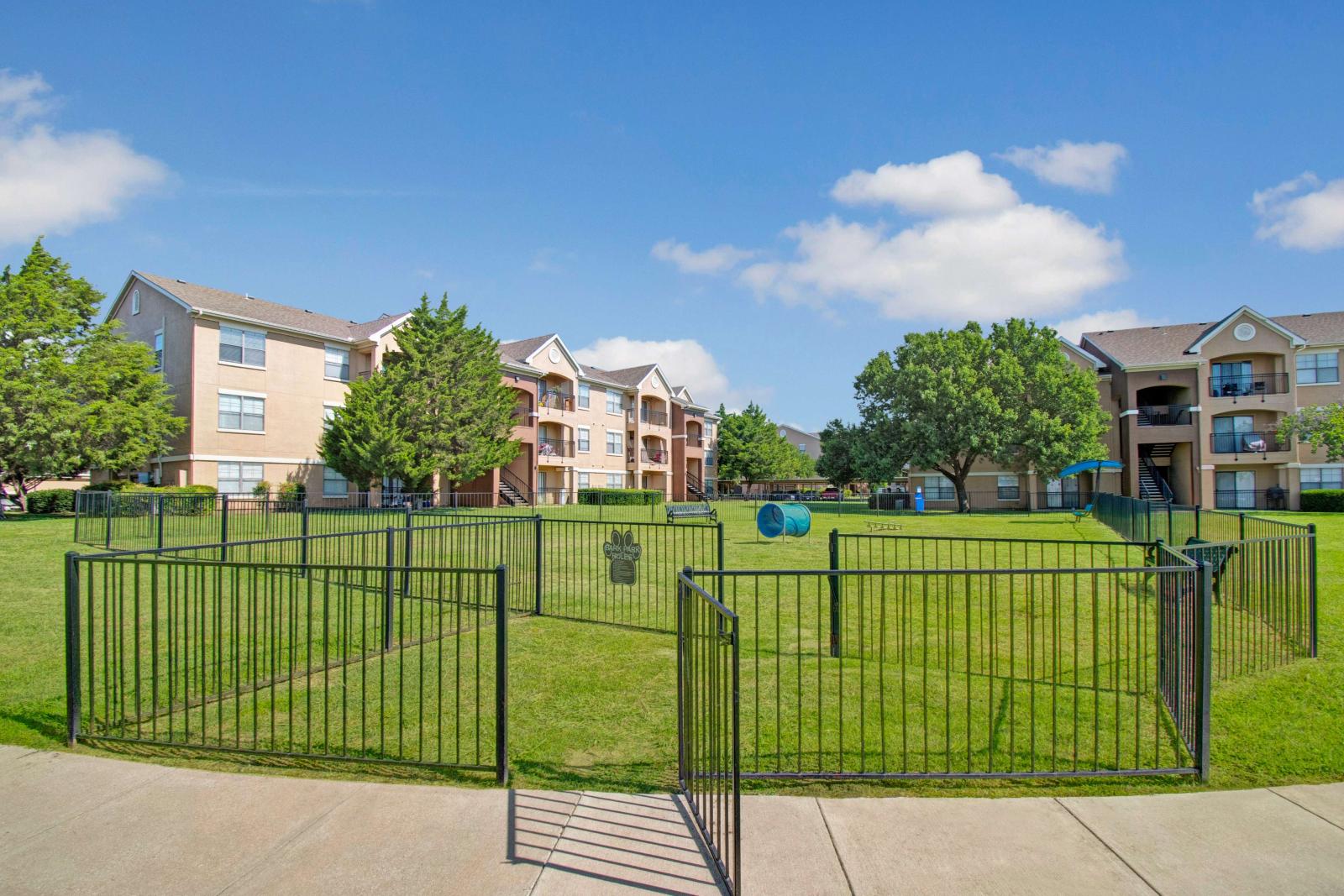Gated entrance to a spacious dog park with agility tunnel and hurdles, surrounded by grassy lawns and apartment buildings.