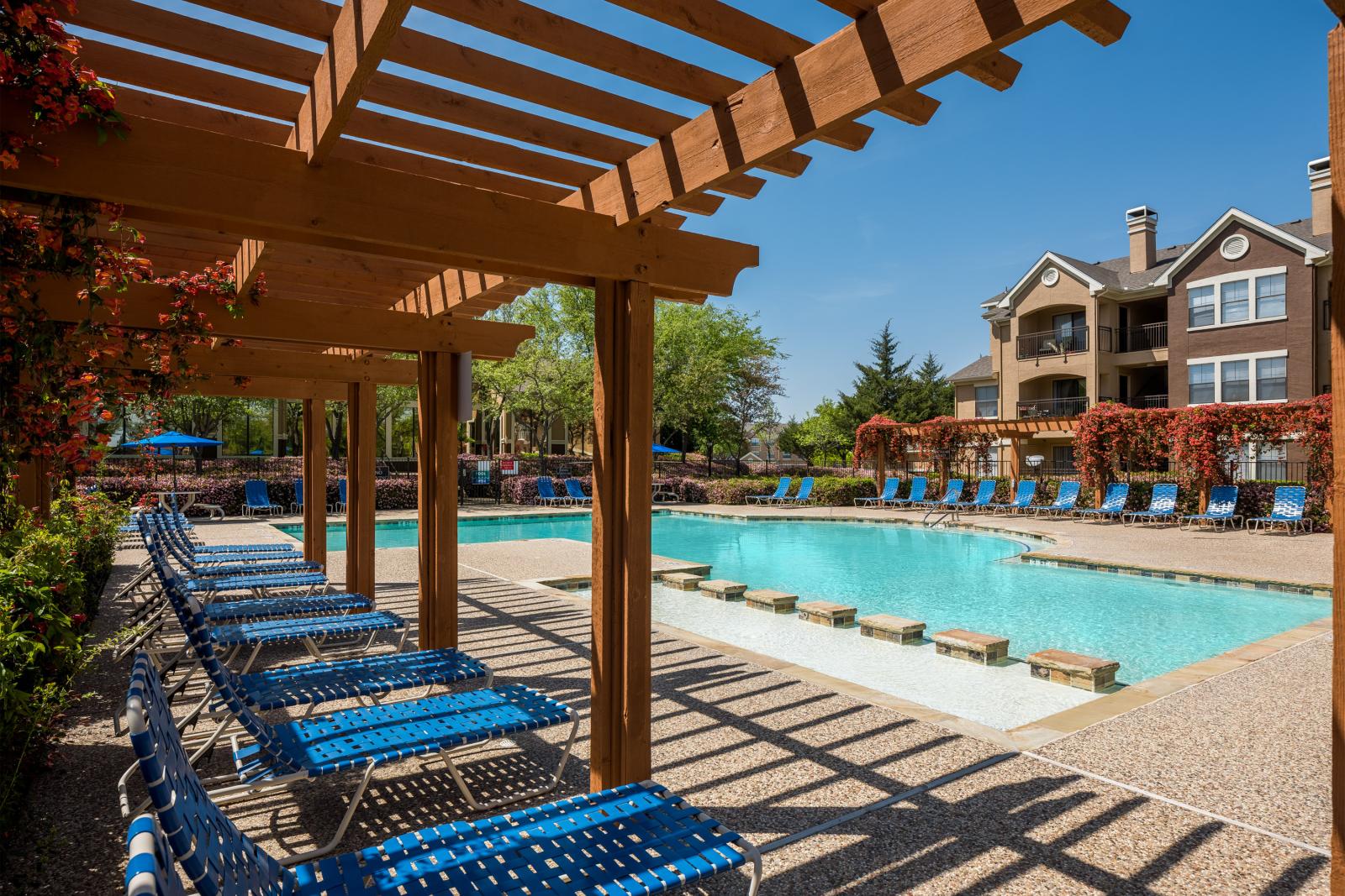 Resort-style swimming pool with stepping stones, surrounded by blue lounge chairs and wooden pergolas draped in vibrant flowering vines, adjacent to a multi-story apartment building.