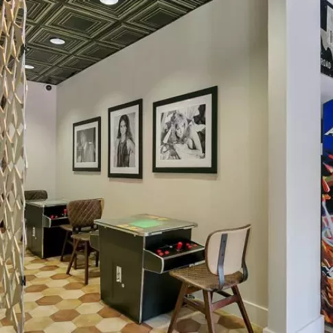 A retro-inspired game room featuring arcade machines, tabletop games, modern chairs, and framed black-and-white portraits on the wall.