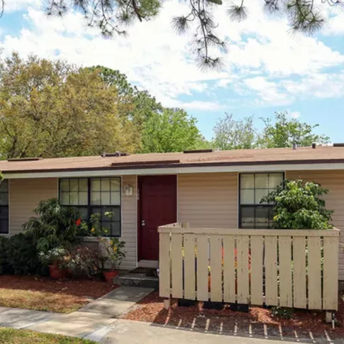 Beige home with a red door, white picket-style fencing, and lush landscaping.