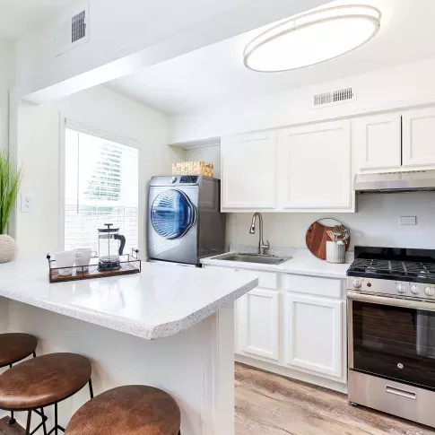 Bright galley-style kitchen with white cabinets, stainless steel appliances, and a stacked washer and dryer next to a breakfast bar with three stools.