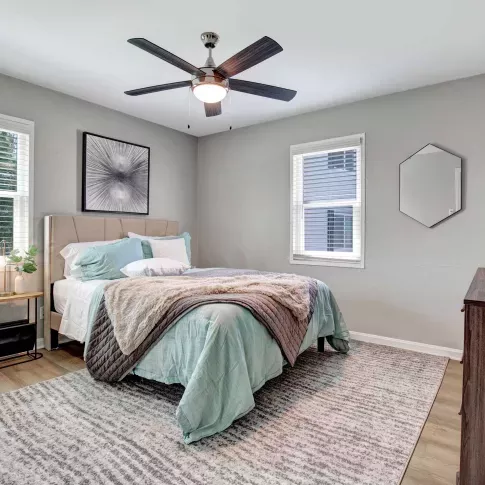 Modern bedroom with wood-style flooring, neutral walls, and a ceiling fan above a queen-sized bed with blue and white bedding.