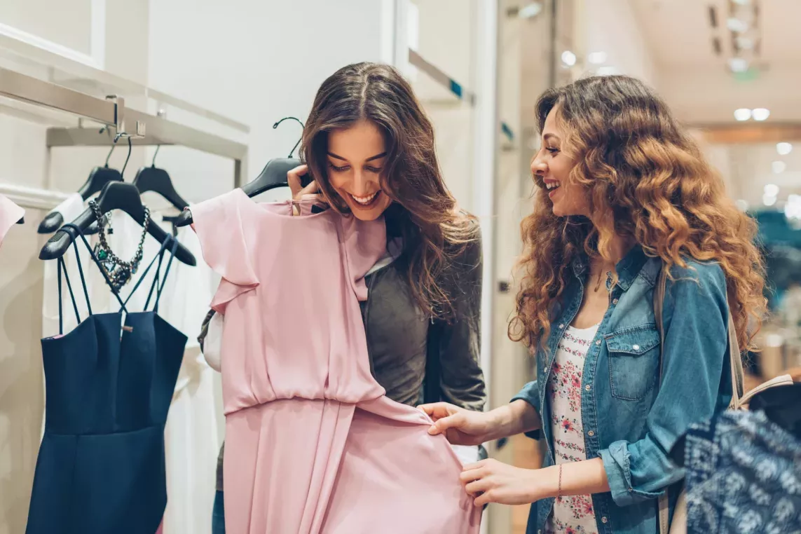 Two women joyfully shopping for clothes, with one holding a light pink dress while the other admires it.