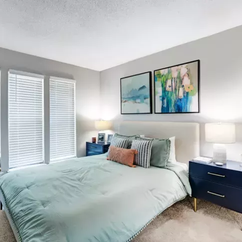 A bright bedroom featuring a neatly made bed with pastel bedding, modern navy nightstands, and abstract wall art above.