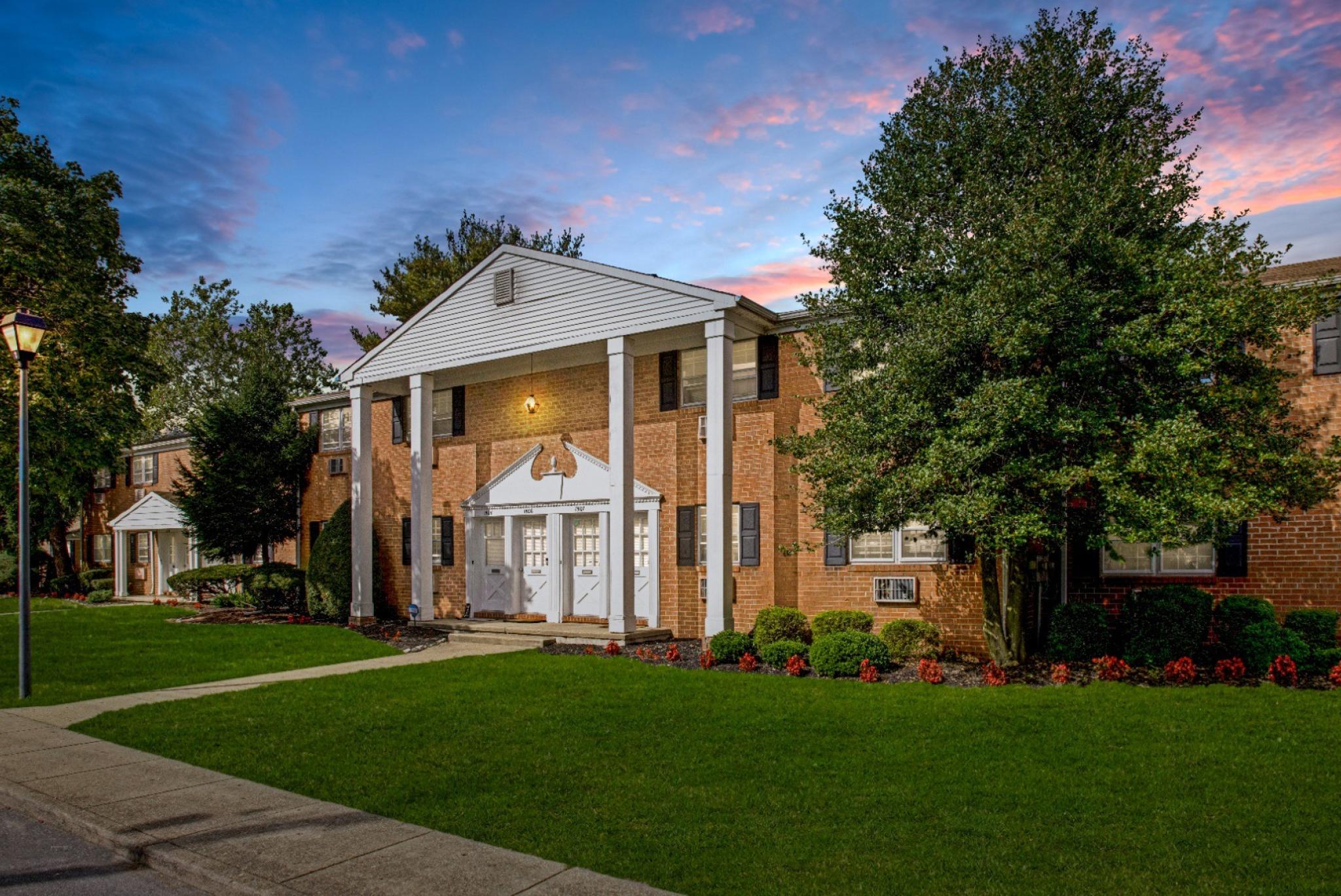 A grand entrance to Silver Lake Manor Apartments at twilight, showcasing a stately red brick building with tall white columns and an elegant gabled roof. The manicured lawn and vibrant flower beds add to the curb appeal, while a lamppost lights up the scenic sidewalk leading to the entrance. The sunset sky in the background creates a warm, inviting ambiance. ​​
