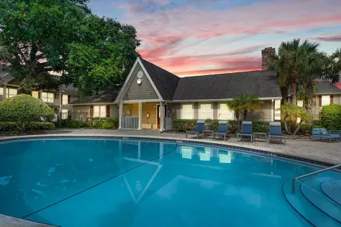 A serene pool area with lounge chairs and lush landscaping, set against a vibrant sunset sky and a cozy clubhouse.