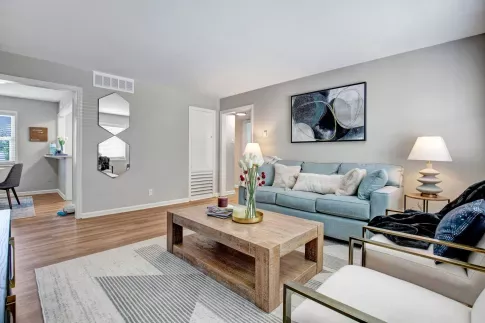 A stylish and cozy living room at Alcove at Seahurst, featuring a soft blue sofa adorned with decorative pillows, a rustic wooden coffee table with a floral centerpiece, and two modern accent chairs with gold metal frames. The space is illuminated by warm table lamps, and abstract wall art enhances the contemporary decor. An open doorway leads to a small dining area, and large windows bring in natural light, creating an inviting atmosphere. ​​