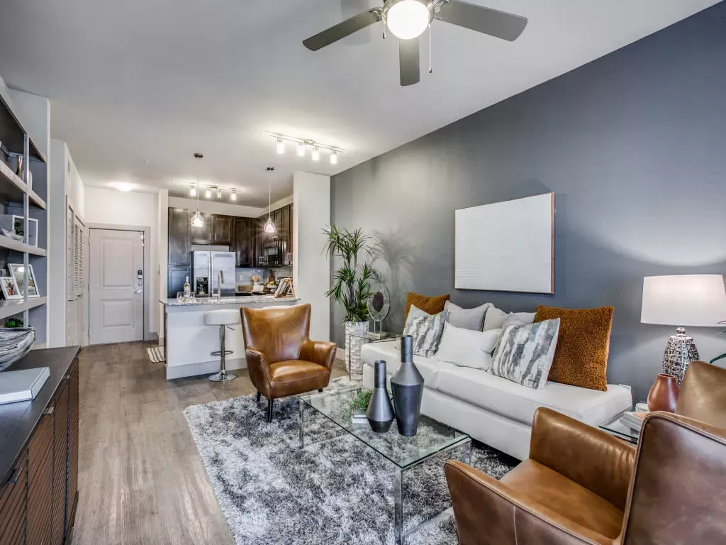 A stylish living room with a white sofa, brown leather chairs, a shag rug, and an open-concept kitchen in the background.