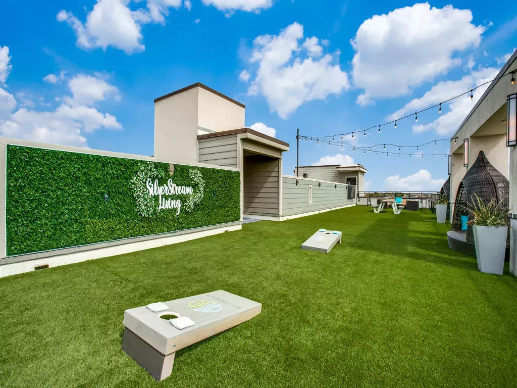 A rooftop lounge area with artificial turf, cornhole boards, string lights, and seating, featuring a "SilverStream Living" sign on a greenery wall.