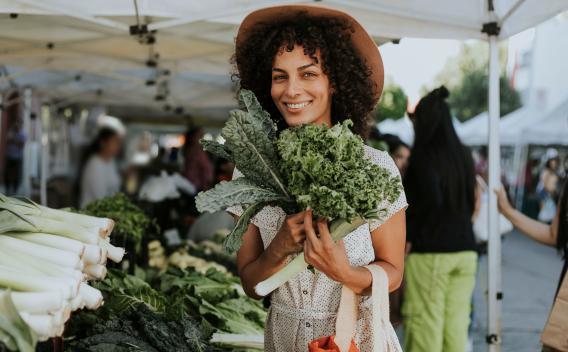 A woman smiles while holding fresh greens at a vibrant outdoor farmers' market.