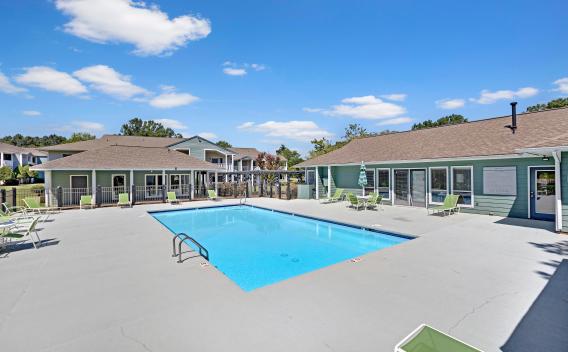 An outdoor swimming pool surrounded by green lounge chairs and a light blue clubhouse under a bright blue sky with fluffy white clouds.