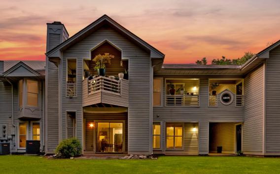 A two-story residential building with balconies, surrounded by greenery, set against a vibrant sunset sky.