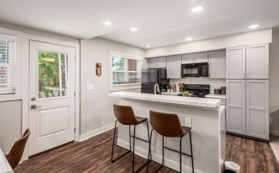 A modern kitchen with gray cabinetry, a marble countertop breakfast bar, and bar stools, complemented by hardwood floors and ample natural light from large windows.
