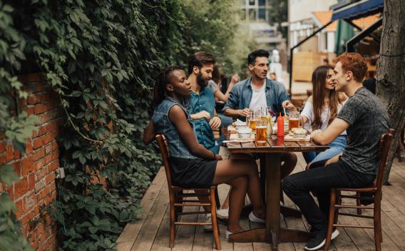 A group of friends enjoys food and drinks together at an outdoor restaurant patio surrounded by greenery.