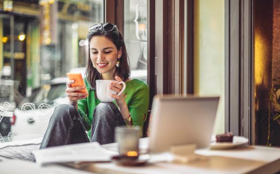 A woman sitting by a cafe window, smiling as she looks at her phone while holding a coffee mug in a cozy and relaxed setting.