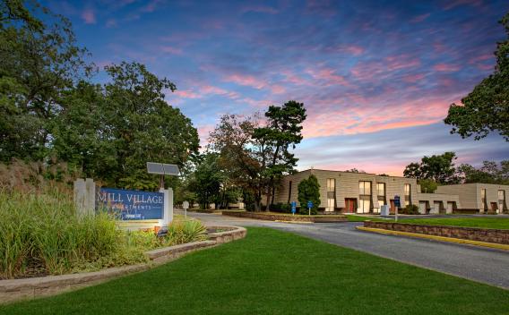 A welcoming entrance to Mill Village Apartments with lush greenery and a vibrant sunset sky.