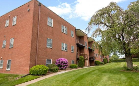 A red brick apartment building with well-maintained green lawns, colorful bushes, and a large tree in the foreground under a partly cloudy sky.