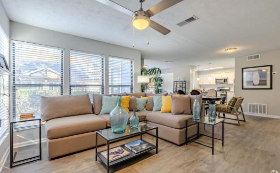 A spacious living room featuring a large sectional sofa with colorful throw pillows, a ceiling fan, and large windows letting in ample natural light.