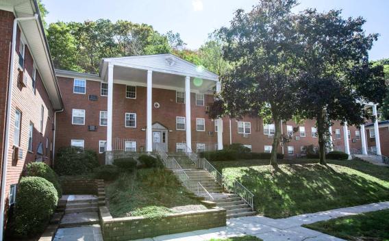 A charming brick apartment building at The Manors Apartments, surrounded by lush greenery and featuring classic white columns at the entrance, offering a welcoming and serene living environment.
