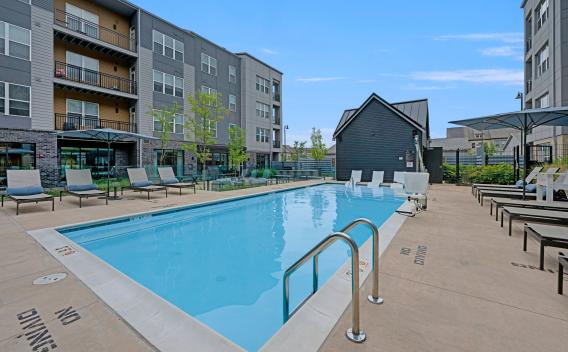 A sparkling swimming pool with poolside lounge chairs, shaded umbrellas, and contemporary apartment buildings in the background, offering a relaxing oasis for residents.