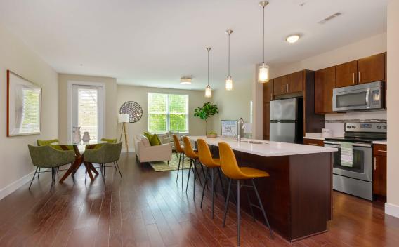 A spacious kitchen and living area at Riverbend at the Charles Luxury Apartments featuring an island with modern pendant lighting, yellow bar stools, and a comfortable seating area bathed in natural light.