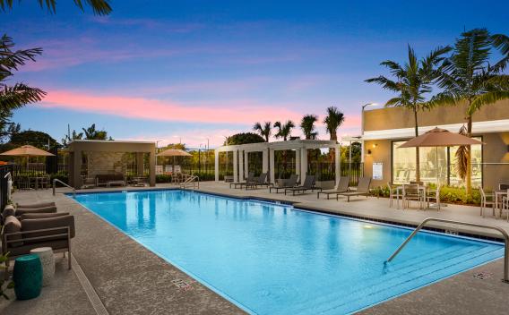 A beautifully lit outdoor swimming pool area at sunset, complete with lounge chairs, cabanas, and lush palm trees for a resort-style experience.