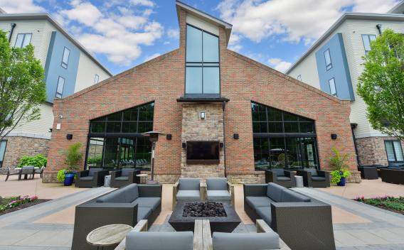 Modern outdoor lounge area featuring a brick and stone clubhouse facade, cozy fire pit, and stylish gray seating surrounded by lush greenery.