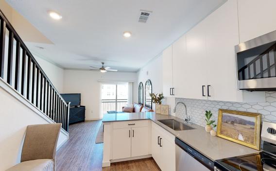 A modern open-concept kitchen with white cabinets, a hexagonal tile backsplash, stainless steel appliances, and a view of a staircase leading to an upper level.