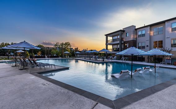 A breathtaking view of The Warner Apartments' pool at sunset, with in-water loungers, striped umbrellas, and elegant lighting creating a serene ambiance.