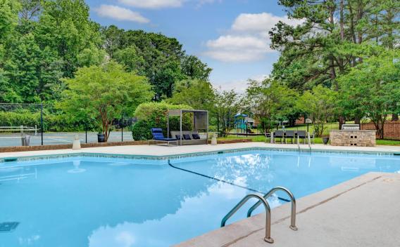 A serene outdoor pool area at Aria North Hills, surrounded by lush greenery and tall trees. The pool features stainless steel handrails, lounge chairs, and a shaded cabana area for relaxation. A tennis court and a children's playground can be seen in the background, offering a variety of recreational amenities. ​​