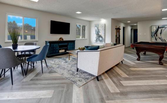 A stylish community lounge with a modern design, featuring a light beige sofa with blue accent pillows, a dark wood pool table with orange tassels, and a round dining table with blue chairs. The room includes a sleek entertainment center with a mounted flat-screen TV, large windows allowing natural light, and abstract art pieces on the walls. The flooring is arranged in a chevron pattern with shades of gray and beige. ​​