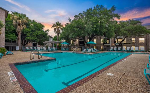 Resort-style swimming pool surrounded by tall palm trees, mature shade trees, and apartment buildings, with plenty of teal lounge chairs and umbrella-covered seating areas on a textured pool deck at sunset.