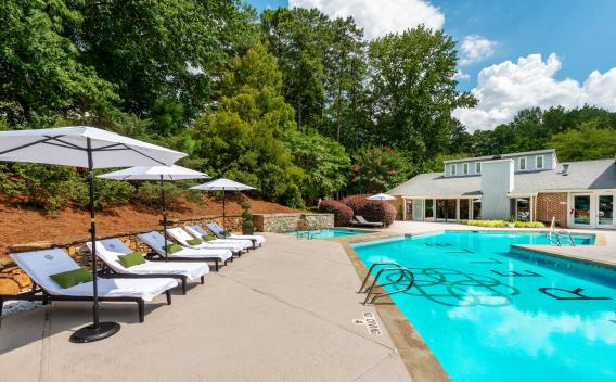 Inviting outdoor pool area with cushioned lounge chairs and white umbrellas, surrounded by lush landscaping and a clubhouse with large windows, offering a tranquil and stylish retreat.