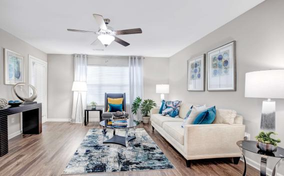 Bright and stylish living room with wood-style flooring, modern decor, a neutral-toned sofa with colorful accent pillows, and a patterned area rug, complemented by ample natural light through large windows.