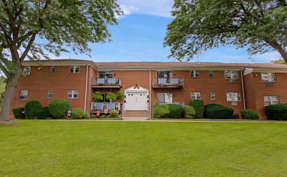 Charming brick apartment building with balconies and well-manicured landscaping, set against a backdrop of mature trees and a wide, green lawn under a clear blue sky.