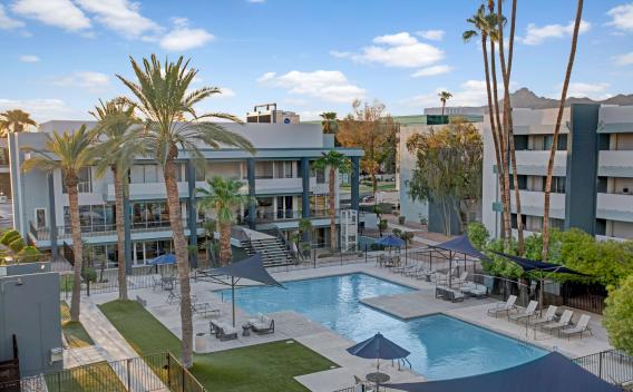 Resort-style pool area surrounded by modern apartment buildings and tall palm trees, featuring two swimming pools, shaded lounge areas with umbrellas and sails, and scenic mountain views in the background.
