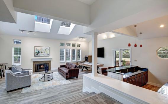 A spacious and modern clubhouse at Chandlers Bay featuring an open-concept design with high ceilings and skylights. The room is furnished with a brown leather sofa, gray armchairs, and a contemporary fireplace with a white mantel. A stylish wooden pool table is positioned near a wall-mounted TV, and a built-in bar area with granite countertops and red pendant lights adds to the upscale ambiance. Large windows and glass doors allow natural light to fill the space, offering views of the outdoor greenery. ​​